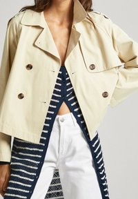 Chaqueta beige de doble botonadura con grandes botones, diseño corto y cuello ancho, superpuesta sobre una blusa a rayas en azul marino y blanco y pantalones blancos.