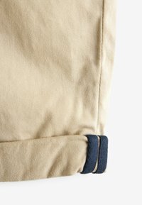 Orlo del pantalone in tessuto beige con una striscia blu navy, che mostra una trama liscia e un design arrotolato. Cuciture visibili lungo la cucitura.
