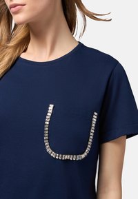 Camiseta de manga corta azul marino con un bolsillo texturizado y adornos plateados en forma de U en la parte delantera. Material de algodón suave.