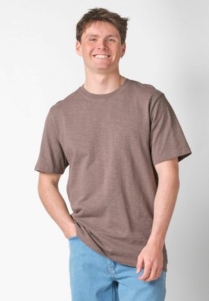 Ung mand smiler, iført en brun kortærmet t-shirt og lyseblå jeans, med den ene hånd i lommen mod en ensfarvet baggrund.