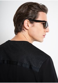 Camisa negra de manga corta con un diseño geométrico texturizado en la parte superior de la espalda y tela suave abajo. Gafas de sol negras complementan el aspecto general.