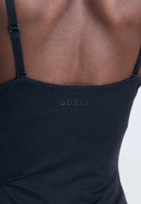 Tuvumā redzams personas augšējais muguras daļa, kas valkā melnu bezpiedurkņu topu ar plānām siksnām, uz kura ir uzdruka "GUESS".