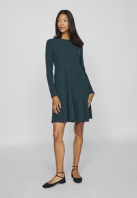 Langärmliges, strukturierte Kleid in dunklem Türkis mit ausgestelltem Saum. Der strukturierte Stoff, der runde Ausschnitt und das figurbetonte Oberteil betonen die Silhouette.