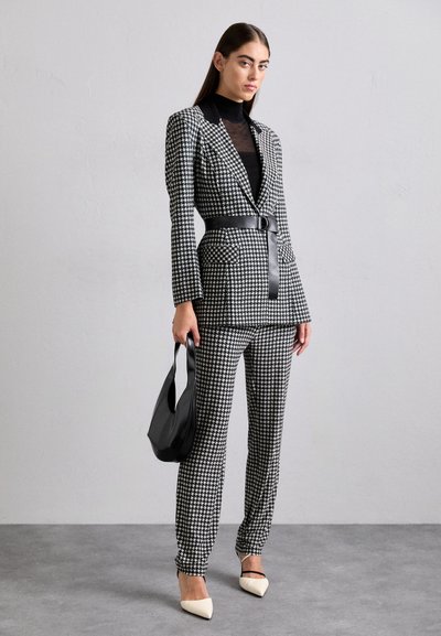 Szürke és fekete houndstooth öltöny, testre szabott blézert hordva, övvel kiemelt derékkal és karcsú fazonú nadrággal, fekete táskával és krémszínű, hegyes orrú magassarkúval párosítva.