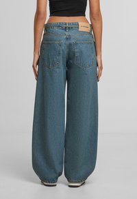 Žena u širokim plavim trapericama i crnom kratkom topu, stoji s leđima okrenutim prema naprijed na jednobojnoj pozadini.