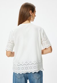 Camiseta blanca con detalles de encaje recortados en las mangas y el dobladillo. Tejido suave, mangas cortas y escote redondeado. Pantalones de mezclilla visibles.