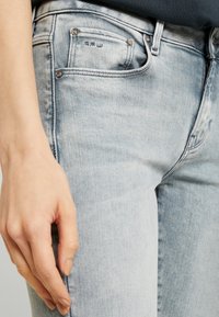 Ljusblå denimjeans med ett blekningseffekt, som har en framlomme, metallnitar och enkla sömnadsdetaljer. Handen placerad på höften.