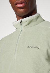 Pull-over en polaire vert clair avec un col montant, une fermeture éclair mi-longue et un logo brodé sur la poitrine. Texture douce, design structuré.