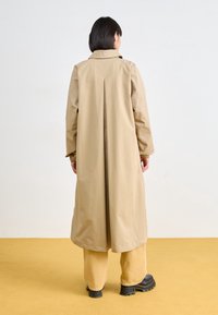 Dlouhý béžový kabát s rozparkem na zádech, límcový výstřih a elastické manžety, v kombinaci se světle žlutými kalhotami a černými platformními botami.