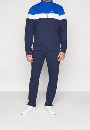 Homme portant un survêtement zippé bleu, blanc et marine avec des baskets blanches, debout devant un fond clair uni.