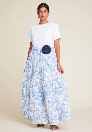 Hvit t-skjorte sammen med et langt, lagdelt blått og hvitt blomstermønster-skjørt med en denimblomst-detalj i midjen og flate sandaler.