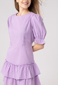 Vestito viola con maniche a sbuffo e scollatura rotonda, arricchito da dettagli in pizzo e un orlo a strati con ruches. Tessuto morbido con una texture liscia.