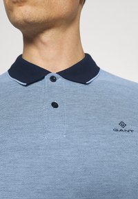 Polo bleu clair avec un tissu texturé, col marine, deux boutons noirs, et un petit logo GANT sur la poitrine gauche.