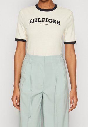 Kvinde iført en cremefarvet t-shirt med sort kant og teksten "HILFIGER", kombineret med højtaljede lyseblå plisserede bukser.