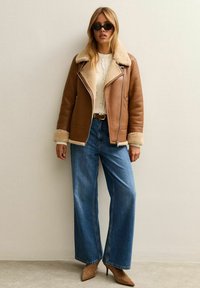 Giacca di pelle marrone con colletto in shearling, abbinata a un maglione bianco sporco, jeans in denim blu a gamba larga e scarpe col tacco beige.