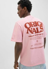 Roosa puuvillane T-särk, mille seljal on julge punane tekst, sealhulgas "ORIGINALS" ja "URBAN AESTHETICS", lõdva istuvuse ja lühikeste varrukatega.
