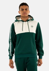 Sudadera con cremallera en verde y blanco, con un bolsillo frontal, que presenta un logo, capucha con cordones y franjas contrastantes en las mangas. Textura de tejido suave.
