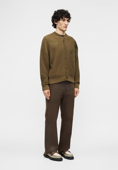 Άνδρας με κατσαρά μαλλιά, φορώντας μια καφέ υφασμάτινη ζακέτα με κουμπιά, καφέ παντελόνια και μπεζ-καφέ παπούτσια, στέκεται μπροστά σε ένα απλό φόντο.