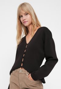 Zwart gebreide trui met een V-hals, knopen aan de voorkant en lange mouwen. Heeft een cropped ontwerp en textielstructuur. Gecombineerd met beige corduroy broek.
