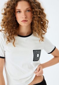 Camiseta blanca de manga corta con cuello de canalé negro y bolsillo, que presenta un patrón bordado geométrico en blanco y negro.