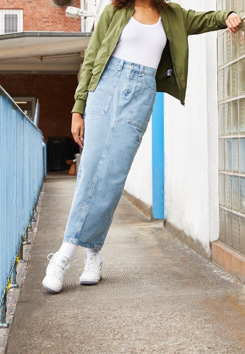 Femme en veste verte, haut blanc, jupe en jean bleu clair et baskets blanches, appuyée contre un mur en blocs de verre sur un passage extérieur.