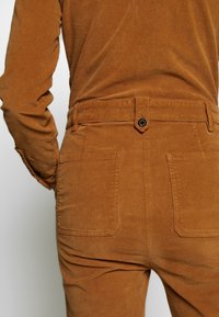 Pantalones de pana marrones con una cintura ajustada, que presentan dos bolsillos traseros, un cierre de botón metálico y una tela suave y texturizada.