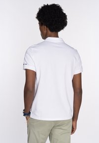 Camisa polo blanca de manga corta, con cuello y textura suave, que presenta un pequeño logo en la manga izquierda. Llevada con pantalones de color claro.
