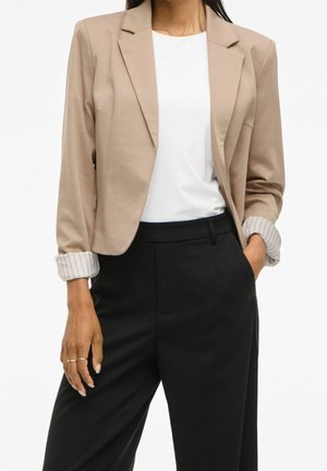 Kobieta w beżowej marynarce z podwiniętymi mankietami w paski, białej bluzce i czarnych spodniach z wysokim stanem, z ręką w kieszeni, na białym tle.