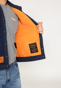 Granatowa kurtka bombowiec z pomarańczowym wnętrzem; ma miękki kołnierz, napy oraz metkę wewnątrz z informacjami o marce i pielęgnacji.