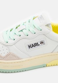 Białe skórzane trampki z miętowymi sznurówkami, żółtą podszewką i jasnobeżowym zamszowym akcentem. Posiadają logo "KARL" z boku.