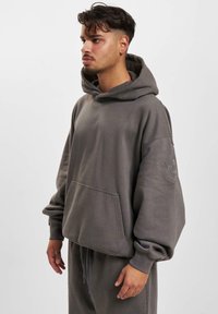 Siva dukserica s kapuljačom s oversized krojem, veliki prednji džep, rebrasti rukavi i izvezeni logo na rukavu. Mekani materijal s suptilnom teksturom.