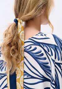 Niebieska i biała bluzka w wzory o swobodnym kroju; zawiera żółtą i granatową wstążkę włożoną we włosy. Miękki materiał.