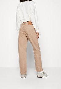 Personne portant un jean beige à jambes droites, un sweat-shirt blanc court, et des baskets blanches, debout devant un fond blanc uni.