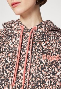 Gros plan d'une personne portant un sweat à capuche noir et rose tacheté avec des cordons roses et le logo "BOSS" brodé sur la poitrine.