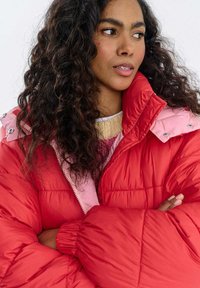 Czerwona i jasnoróżowa kurtka puchowa z wysokim kołnierzem, pikowaną fakturą i zamkiem błyskawicznym. Posiada duże rękawy i srebrne zatrzaski na ramionach.