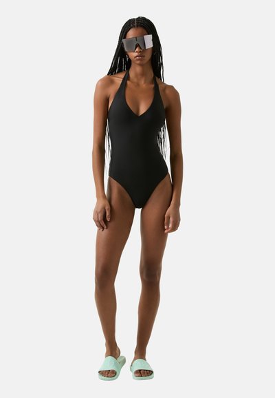 Black halter neck swimsuit with a deep V neckline, paired with mint green slides and large mirrored sunglasses, showcasing a sleek, minimalist design.