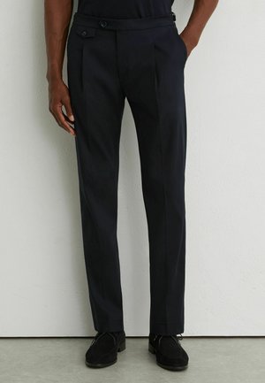 Man wearing black straight-fit dress pants and black shoes, standing against a white wall with hands partially in pockets.