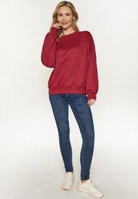 Piros pulóver kapucnival, laza szabással, bordás mandzsettákkal és aljjal. Kék skinny farmernadrággal és fehér sportcipővel párosítva.