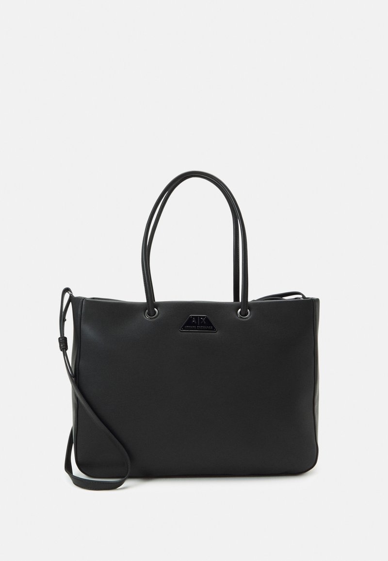 Sac fourre-tout en cuir noir avec doubles poignées, une bandoulière amovible et une petite plaque triangulaire avec logo à l'avant.