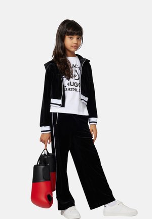 Black velour tracksuit with white stripes, cropped zip-up jacket, and loose pants. White graphic t-shirt underneath. Carries a black and red bag.