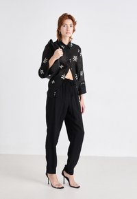 Cămașă neagră transparentă cu butoni, decorată cu modele brodate albe, asortată cu pantaloni negri cu șnur și sandale negre cu toc înalt.