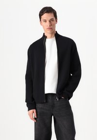Black, zippered jacket with a high collar, made of a smooth fabric. Worn over a white shirt, paired with dark jeans for a minimalist look.