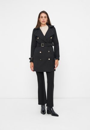 Femme debout portant un trench-coat noir ceinturé avec des boutons dorés, pantalon noir, bottines noires et un haut clair sur fond blanc.