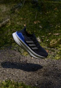 Svart atletisk sko med texturerad tygovandel, vit mellansula och blå accenter. Har tre svarta ränder på sidan. Lättviktig design.