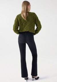 Suéter verde texturizado de cultivo con un patrón geométrico, combinado con jeans ajustados de campana negros. Unos zapatos de tacón alto negros completan el look.