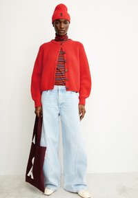 Rode geribde cardigan met knopen over een zwart-rode gestreepte coltrui, lichtblauwe wijde jeans en een kastanjebruine tote bag. Rode beanie met accent.