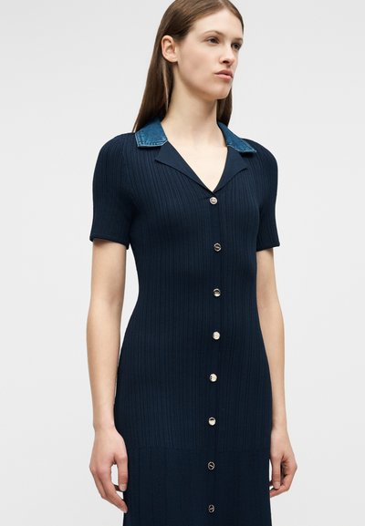 Γυναίκα που φοράει φόρεμα σε στενή πλέξη σε ναυτικό μπλε χρώμα με κοντά μανίκια, ασημένια κουμπιά στην μπροστινή πλευρά και αντιθετικό γιακά από τζιν.