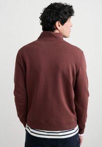 Uomo con capelli ricci scuri che indossa una giacca bordeaux e una camicia a righe, rivolto di lato su uno sfondo chiaro e semplice.