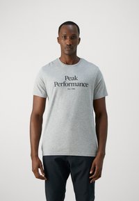 Grå t-skjorte laget av bomull, med korte ermer og rund hals. Fronten har svart tekst "Peak Performance Est. 1986" i en fet skrifttype.