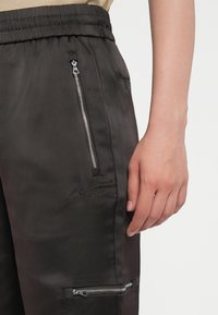 Black satin joggers featuring a drawstring waist, side zippered pockets, embroidered logo, and a sleek, smooth texture.